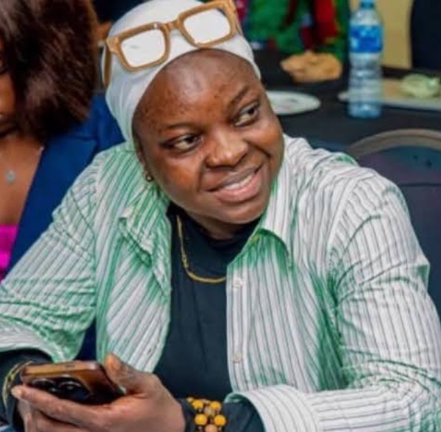 Suliat Yetunde Ogun in a white turban, green stripped shirt with black under shirt, smiling and holding her phone